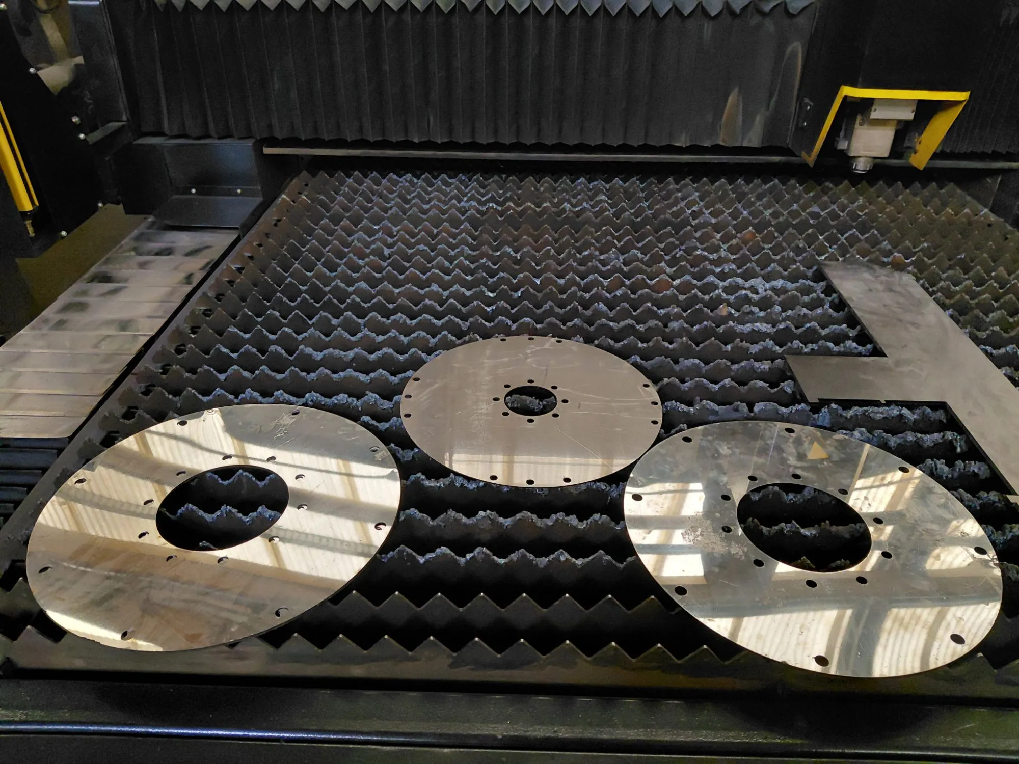 Tres discos metálicos con perforaciones recién cortados sobre la mesa de la máquina láser